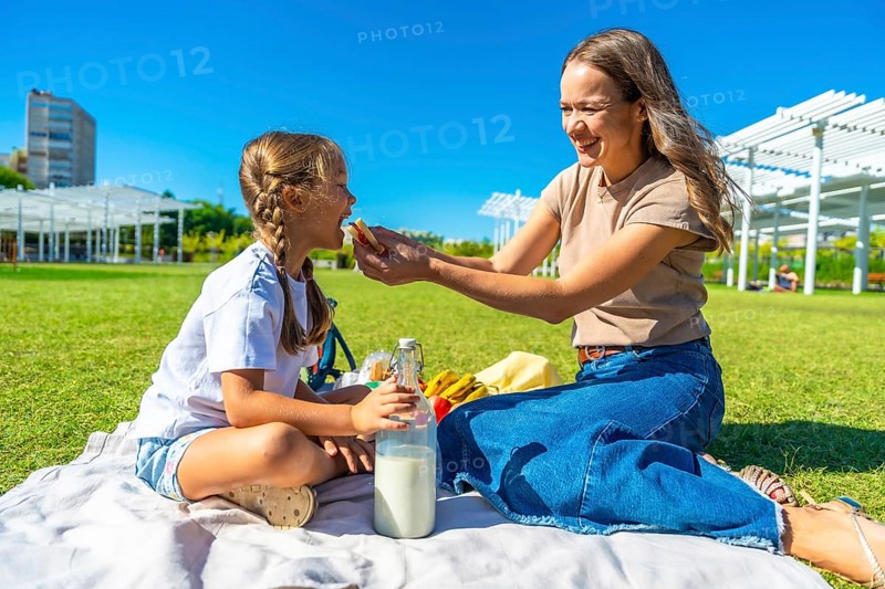 Young mother feeding her little daughter a sandwich during a sunny summer picnic on a blanket in the park, sharing tender moments of bonding, joy and outdoor relaxation