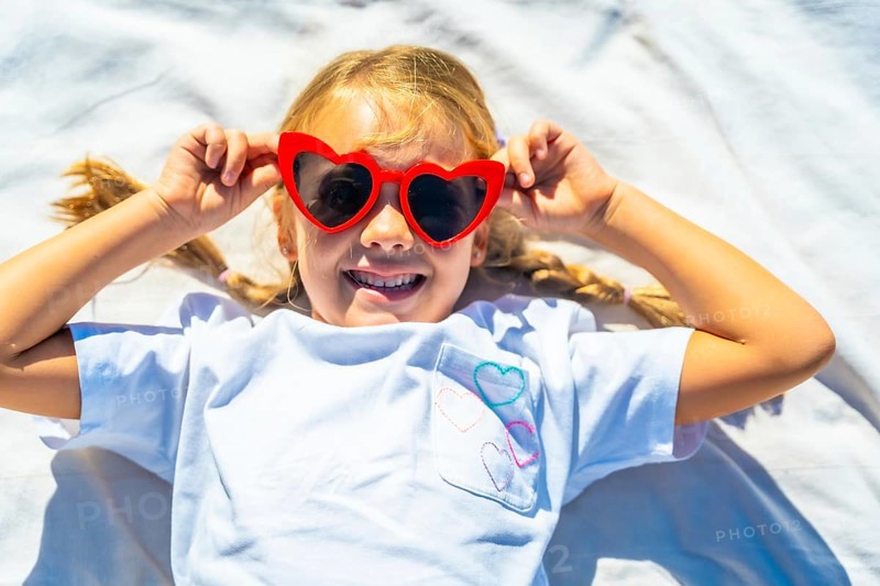 Young child lying outdoors on a white blanket, expressing joy and playing with vibrant red heart shaped sunglasses, embodying summer fun and carefree days