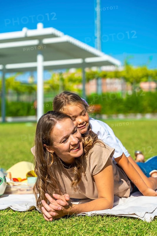 Mother and daughter lying together on a blanket, smiling and hugging, enjoying summer day outdoors in a green park, symbolizing family love, happiness, parenting, and childhood