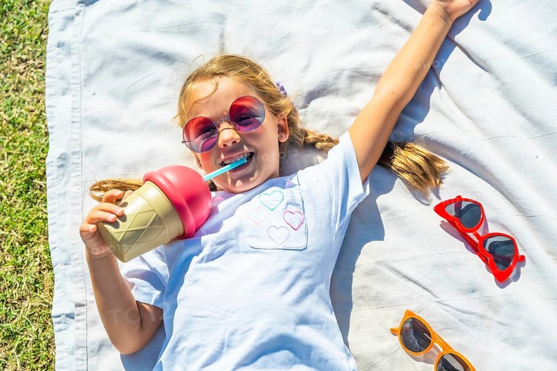 Young girl lying on a white blanket in green grass, smiling while drinking from an ice cream shaped cup through a blue straw, enjoying summer fun outdoors