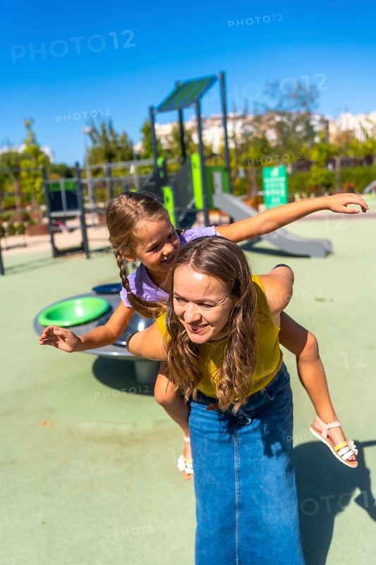 Mother gives daughter a piggyback at a sunny playground, both laughing and smiling as they enjoy playful summer fun, bonding and carefree outdoor family time