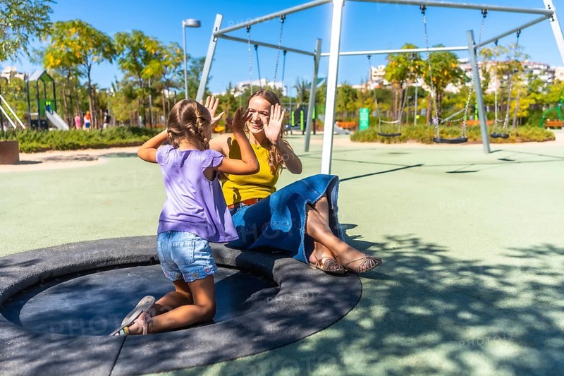 Mother and daughter enjoying playful interaction while sitting on a trampoline in a modern park, creating shared moments of happiness and connection under the sun