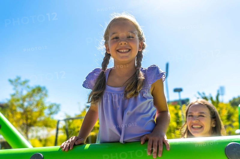 Young girl with pigtails smiling at the camera while her mother watches from playground equipment on a sunny day, enjoying outdoor family fun and carefree childhood joy