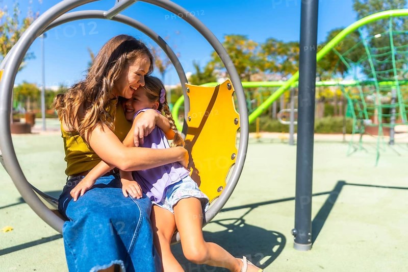 Mother and daughter share a warm hug and bright smiles on a sunny playground, enjoying playful bonding, affection and joyful family time outdoors