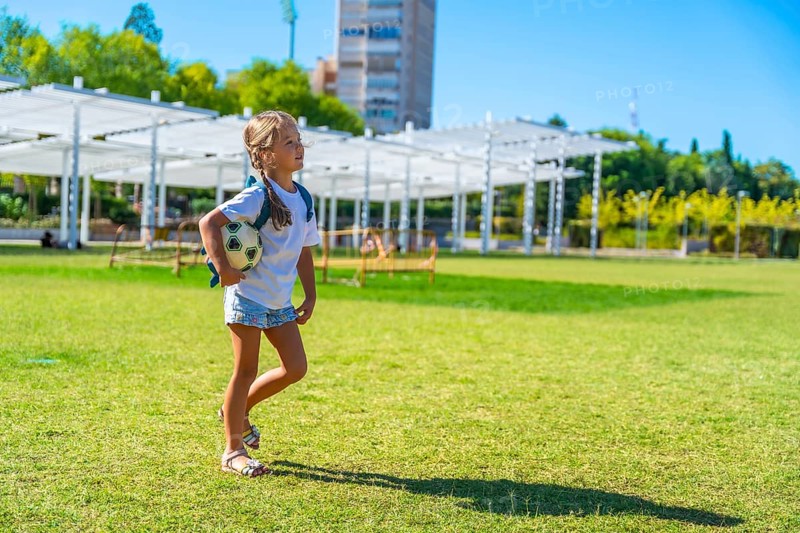 Young girl walking across a sunny green park field, carrying a small soccer ball and backpack, smiling as she enjoys a carefree summer day of play and outdoor fun