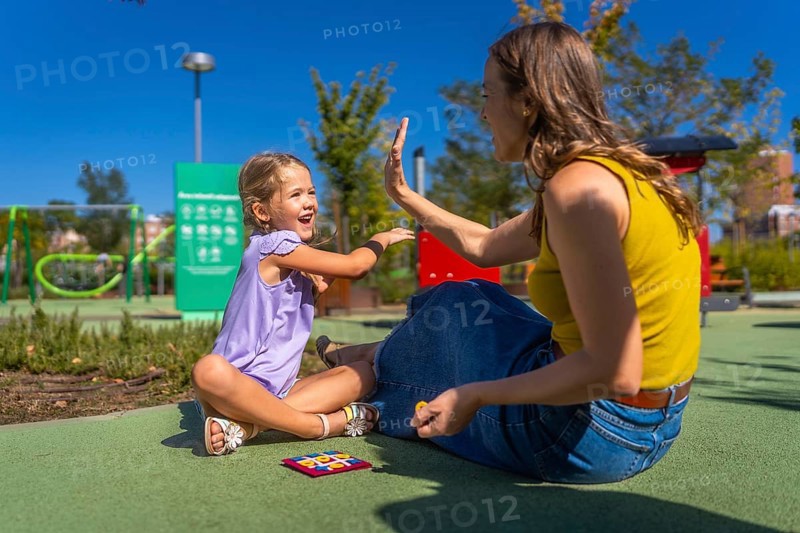Mother and daughter sitting on playground ground, smiling and exchanging a joyful high five while playing a game on a sunny summer day