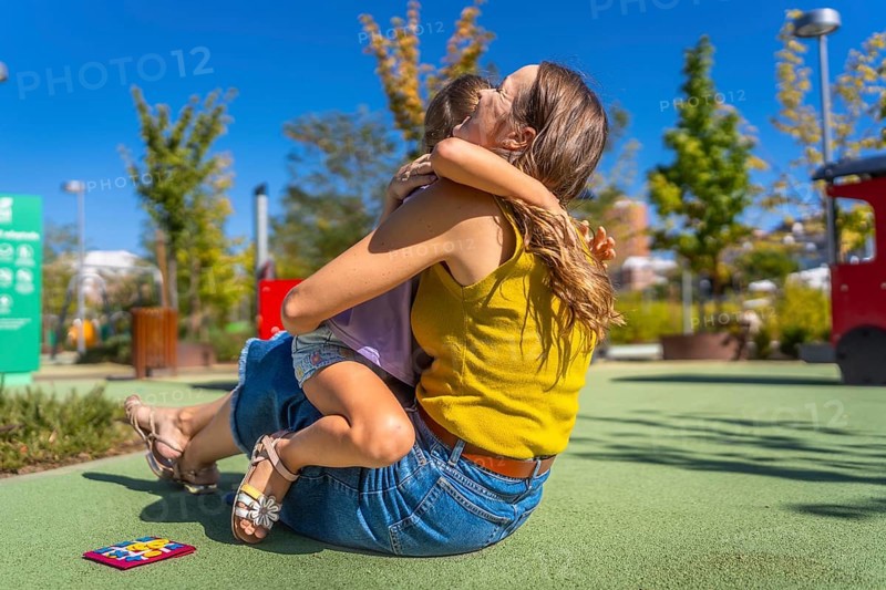 Mother and daughter embracing with affection on a soft ground surface at an outdoor playground, enjoying a moment of connection and family love on a sunny day