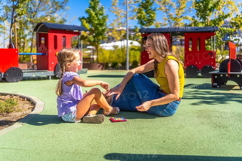 Mother and daughter sitting on soft flooring at a public playground, laughing happily during a sunny day, sharing a bonding moment while playing together