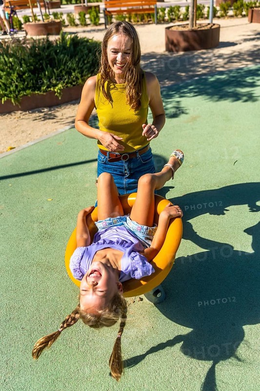 Mother and young daughter share a sunny playground moment as the girl hangs upside down on a spinner, laughing and radiating energetic, carefree family joy and playful bonding