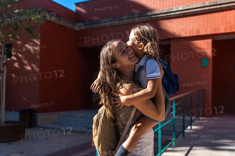 Mother holding her young daughter, both smiling and showing affection, standing outdoors near a school building with backpacks, representing family support and the back to school experience