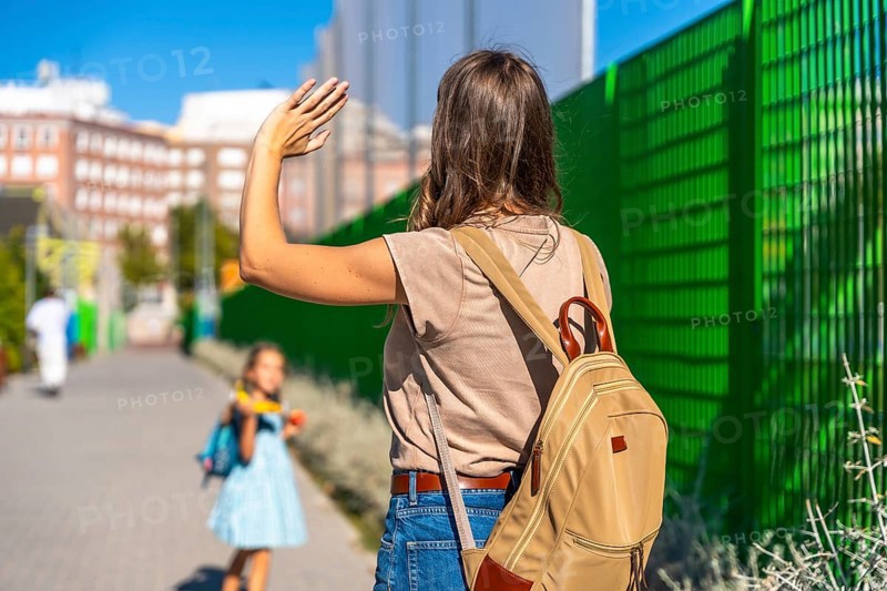 Mother waving goodbye, watching her young daughter with a backpack walk independently towards school, symbolizing back to school, parenting, and family separation emotions