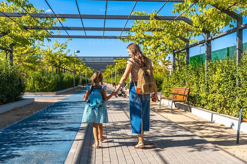 Mother and daughter, wearing backpacks, walking along a sunny pathway lined with green trees and bushes, holding hands on their way to school or kindergarten