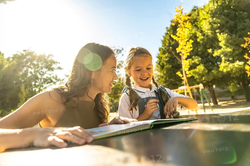Mother and daughter smiling and laughing while reading a book together at an outdoor table in a sunny park or school playground, enjoying warm quality time and learning