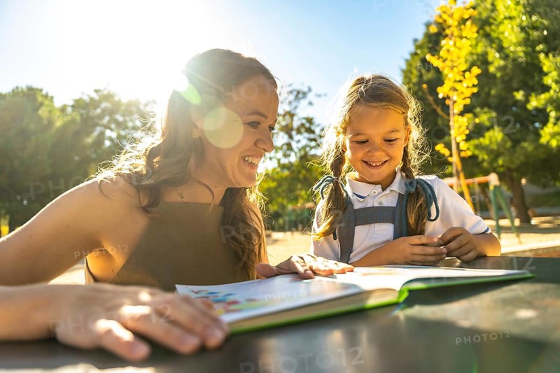 Mother and daughter smiling together at an outdoor schoolyard table, reading a book and bonding over learning in sunshine, casual uniforms and leafy surroundings
