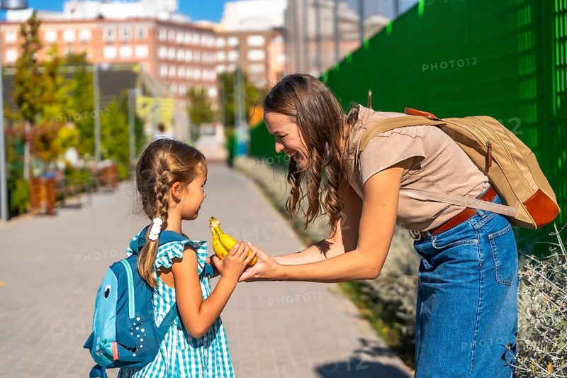 Mother smiles as she hands her schoolgirl a banana on a sunny walk, nurturing healthy habits and bonding before class on an urban neighborhood street