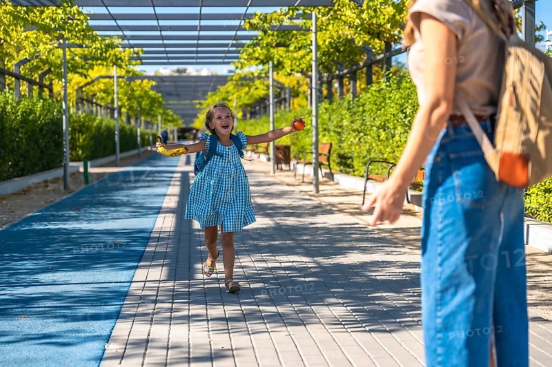 Excited schoolgirl with backpack runs toward her mother in a sunny park, holding a banana after school, joyful reunion and carefree summer energy between parent and child