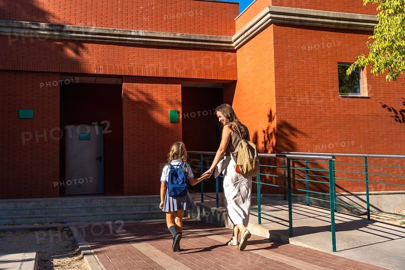 Mother holding hands with her daughter, both walking together towards a brick school building on a sunny day, representing family support and the start of education