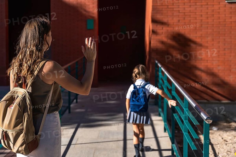 Mother waving goodbye to her elementary age daughter wearing a school uniform and backpack, walking inside a brick school building on a sunny day during drop off