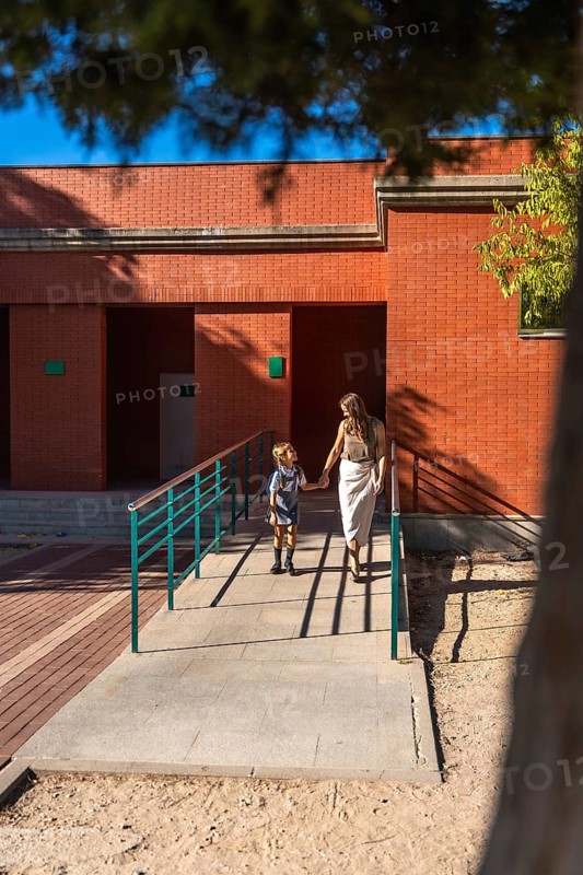 Mother and daughter holding hands and walking together, approaching a school building on a sunny day, symbolizing back to school, education, and family support