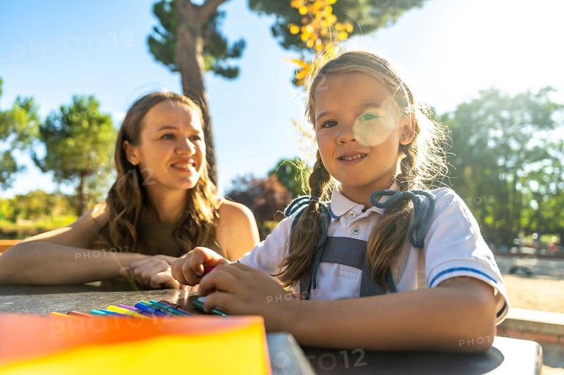 Young daughter and her mother enjoying a sunny day outdoors at the school playground, the child drawing on a table, representing education, family, and childhood