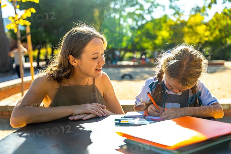 Mother watching her daughter drawing and doing homework at an outdoor table at school, enjoying a moment of family learning and support in the warm sunlight