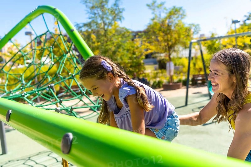 Playful daughter laughing as she climbs a green playground structure while mother smiles and supports her, sharing a joyful outdoor moment of family bonding and carefree summer fun