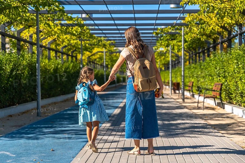 Mother and daughter holding hands and walking together along a paved path, carrying backpacks and heading towards school on a bright sunny day, ready for learning