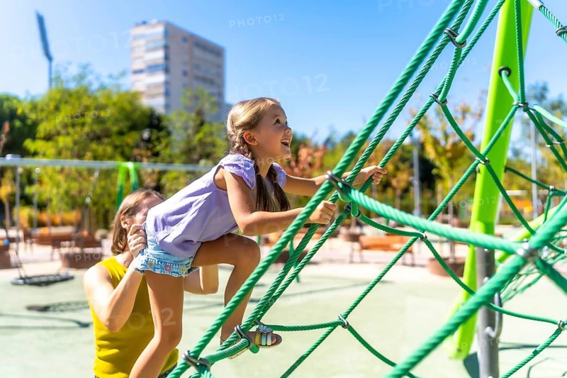 Mother assisting her happy daughter climbing a green rope jungle gym on a sunny day, experiencing childhood fun and family activity together in a park