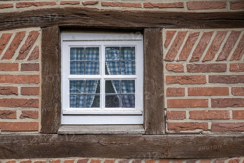 A half-timbered house window with wooden beams and blue checkered curtains, surrounded by red tiles, Münsterland, North Rhine-Westphalia, Germany