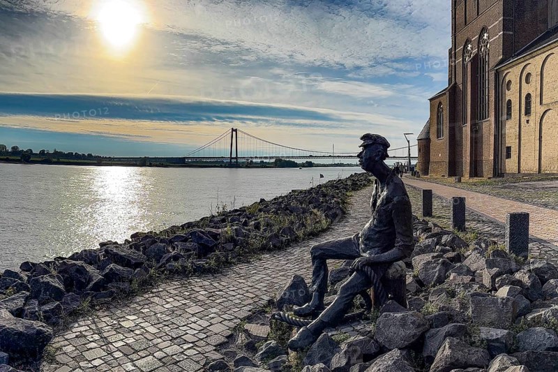 Photo with melancholy mood of bronze sculpture Poortekerl looking at Rhine waiting for ship to unload, Rhine navigation inland shipping, reminiscence of old times, bronze statue by artist Heide Friede-Kinalzik, Emmerich, Lower Rhine, North Rhine-Westphalia, Germany