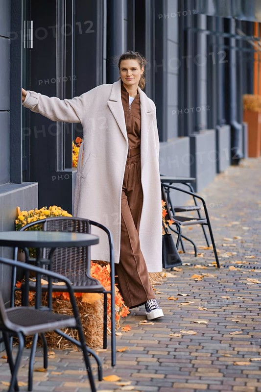 A woman stands near a sidewalk cafe, wearing a trendy outfit with a long coat. The surrounding area features vibrant orange and yellow leaves scattered on the ground, creating an autumn atmosphere