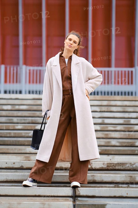 A confident woman stands on the steps wearing a brown tracksuit and a long cream coat. She carries a black bag and smiles, presenting a fashionable look against a contemporary background