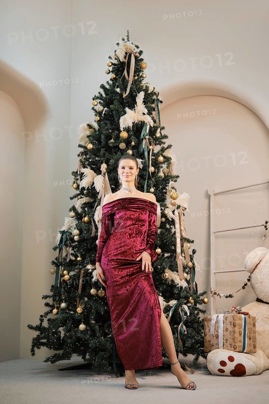 A sophisticated woman stands next to a beautifully decorated Christmas tree adorned with golden ornaments and ribbons. She wears a striking red velvet gown, exuding holiday charm and elegance