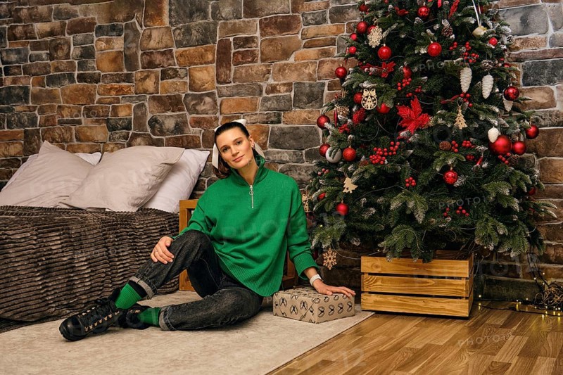 A cheerful woman sits on the floor beside a beautifully decorated Christmas tree in a cozy indoor space. The room features rustic stone walls and festive decorations that create a warm atmosphere