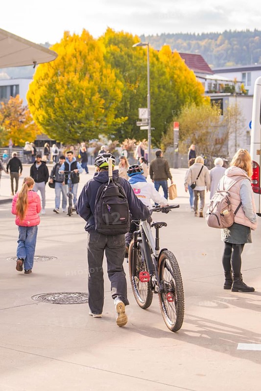A man pushes his bike across a sidewalk while people and autumn trees form the backdrop, Urschelherbst Street Festival, Nagold, Germany