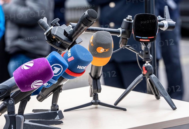 Microphones from various radio stations, television stations, standing on the table, at a press conference