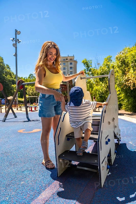 Happy mother assisting her toddler son while he's climbing stairs on a playground structure, enjoying a sunny day at the park