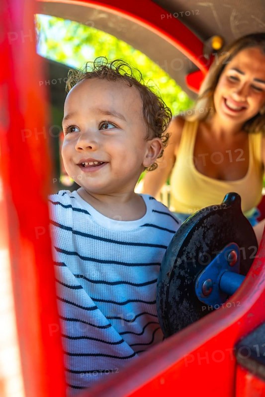 Smiling toddler playing joyfully on a playground slide while mother watches nearby, creating moments of fun and togetherness in the sun