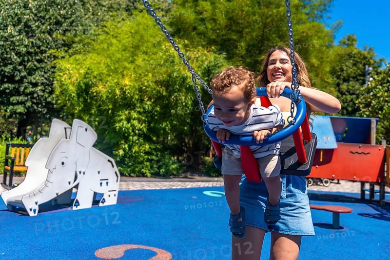 Young mother having fun with her baby son, pushing him on a swing in a colorful playground during a sunny summer day