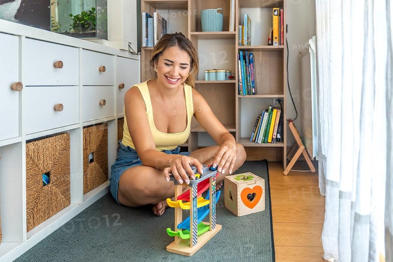 Young woman playing with a toy car ramp and smiling while sitting on a play mat in a child's playroom