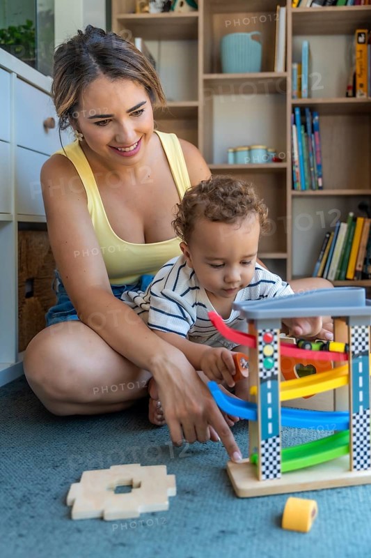Mother and baby son sitting on the floor, joyfully playing together with a colorful toy car and ramp, fostering fun and bonding moments