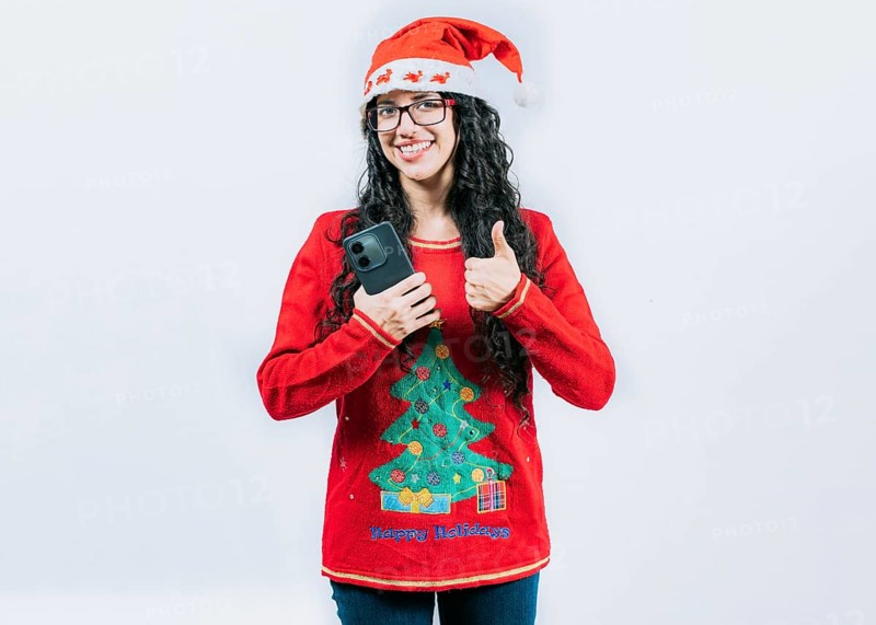 Smiling girl in Christmas hat holding phone with thumb up isolated. Positive young woman in Christmas hat holding smartphone gesturing ok