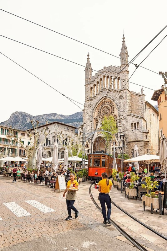 Waiters walking on a square with outdoor restaurants and an old vintage tram driving across the square with lot of tourists, Soller, Mallorca, Spain