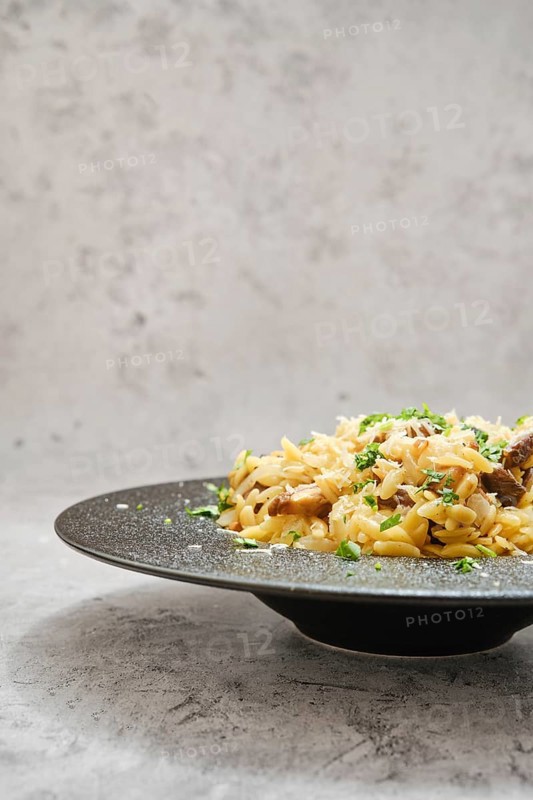 A delicious serving of risoni pasta on a black plate, topped with porcini mushrooms and sprinkled with fresh herbs. The background is neutral, highlighting the dish
