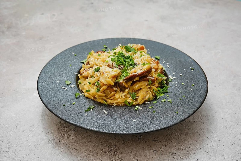 A delicious dish of risoni with porcini mushrooms is served in a sleek black bowl. Fresh herbs and grated cheese garnish the meal, showcasing a blend of flavors ideal for a cozy dinner