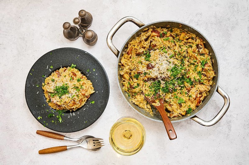 Risoni with porcini mushrooms is elegantly displayed in a pot and on a plate, garnished with herbs and cheese. A glass of wine and seasonings complete the inviting kitchen atmosphere