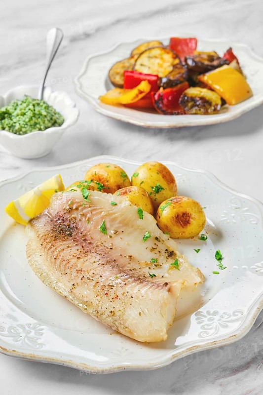Delicious fillet of fish is plated beside roasted vegetables and seasoned baby potatoes. Fresh lemon and a green pesto sauce add vibrant flavors. Carefully prepared for a satisfying meal