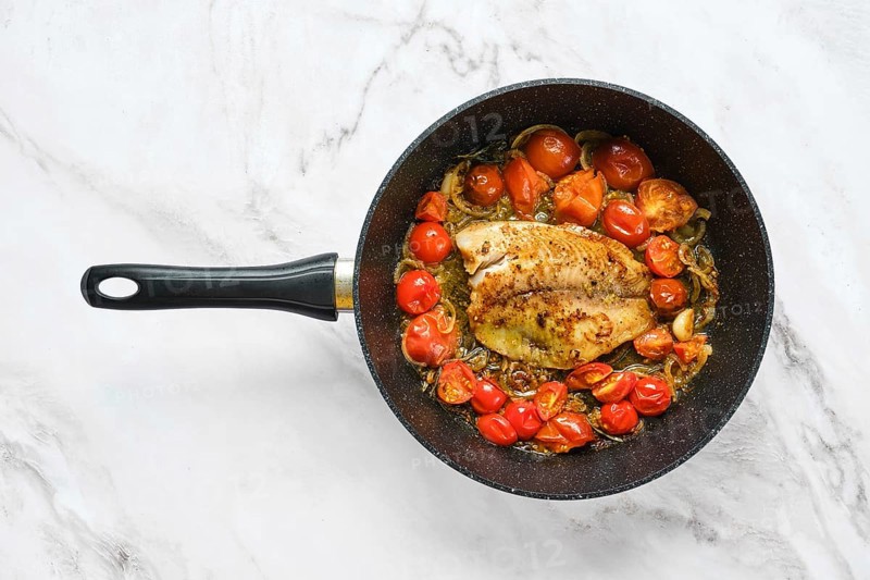 A frying pan holds a seasoned fish fillet surrounded by bright cherry tomatoes. The scene captures a fresh, healthy meal preparation on a modern kitchen counter with marble design
