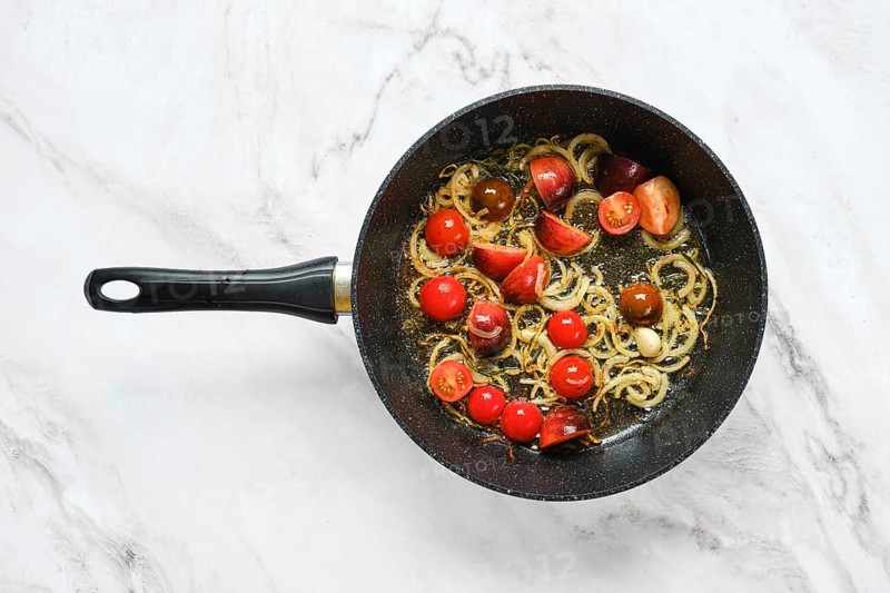 Chopped onions and cherry tomatoes simmer in a black frying pan on a smooth marble surface. The ingredients are seasoned and combined for a flavorful dish in the making, showcasing vibrant colors