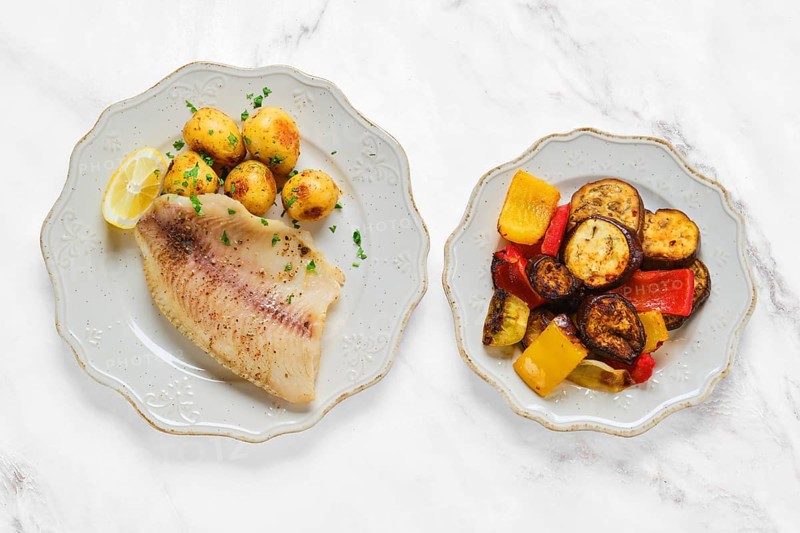 A beautifully plated dish features grilled white fish alongside tender baby potatoes and a colorful mix of roasted vegetables. The meal is garnished with fresh herbs and lemon